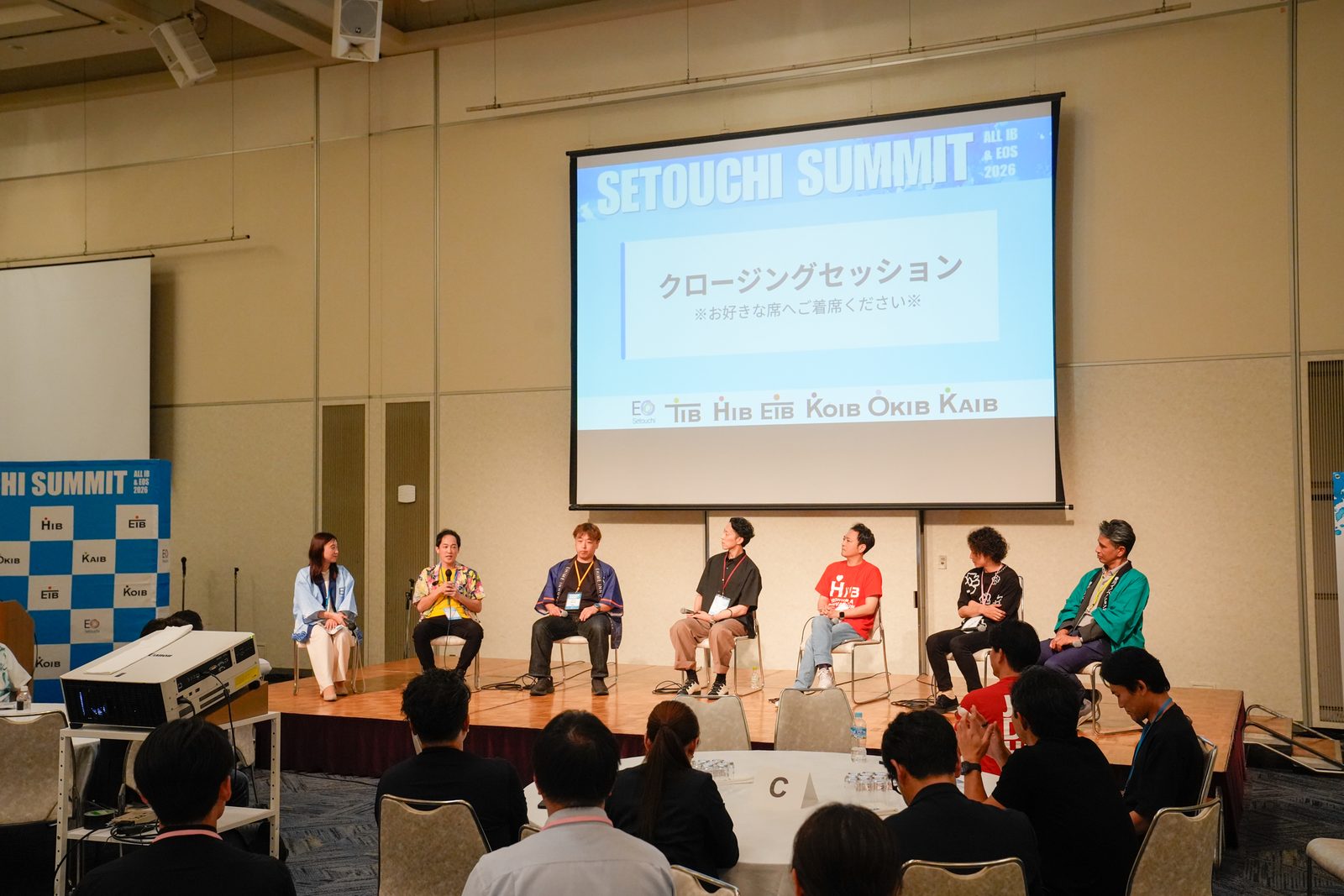 スクリーンに「クロージングセッション」と各地域イノベーションベースのロゴが並び、壇上に7名が座って語り合っている様子