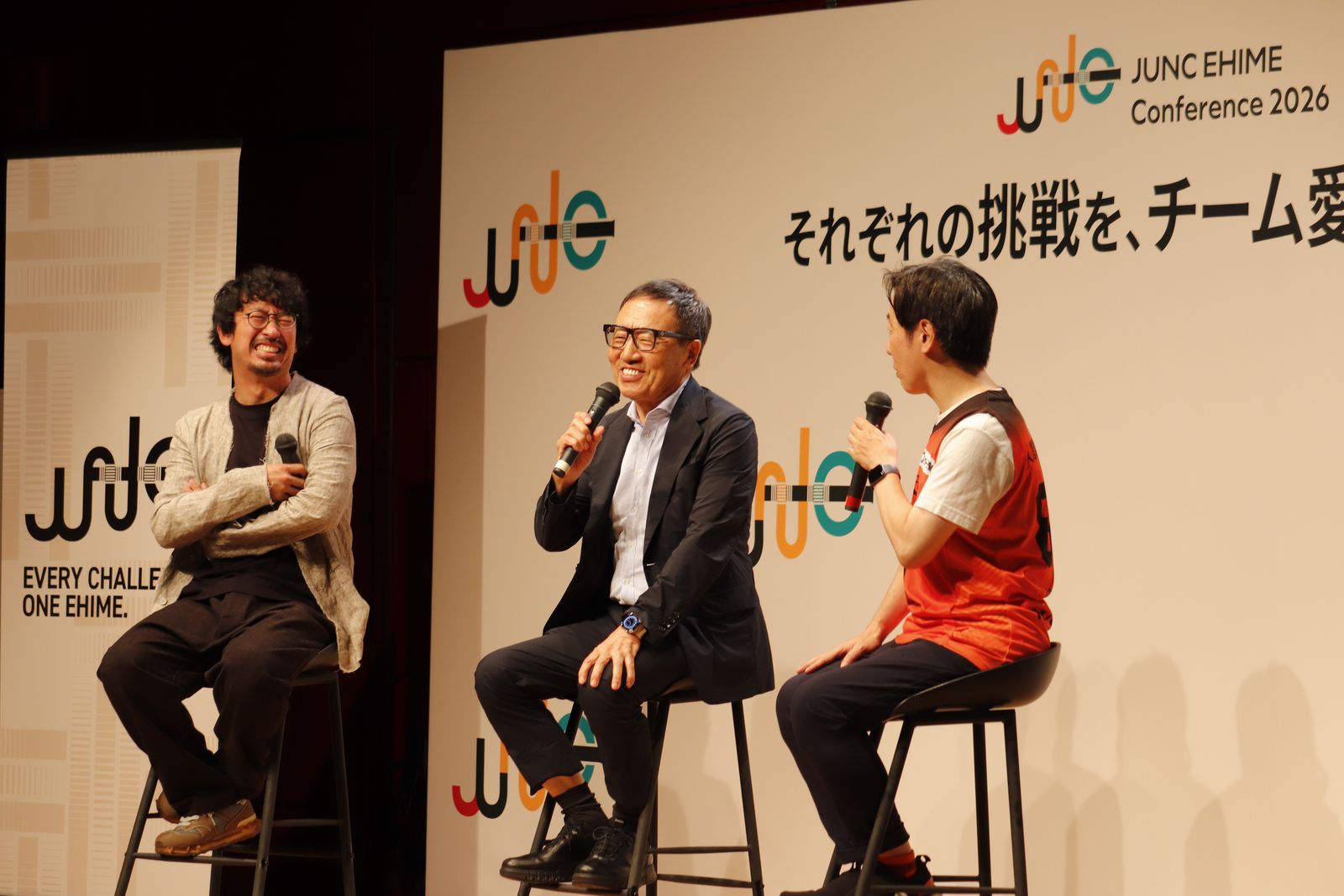 JUNC EHIME Conference 2026のボードの前で、ハイチェアに座って語り合う登壇者3名