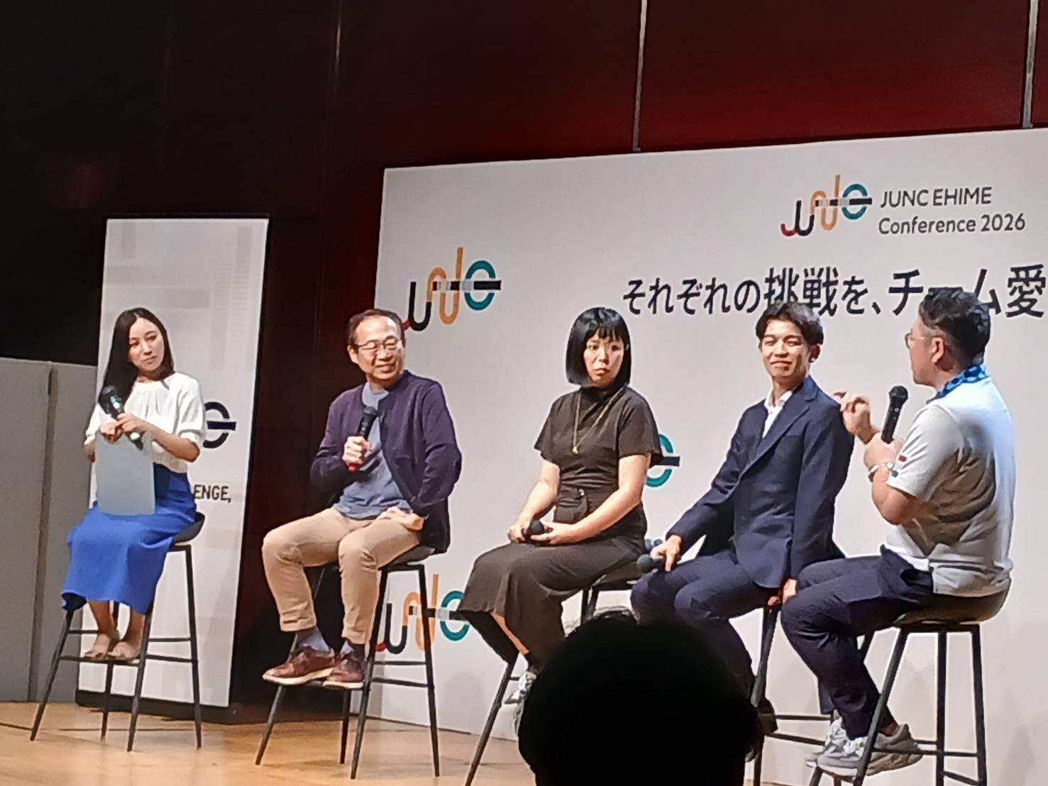 JUNC EHIME Conference 2026のボードの前で、ハイチェアに座る登壇者5名
